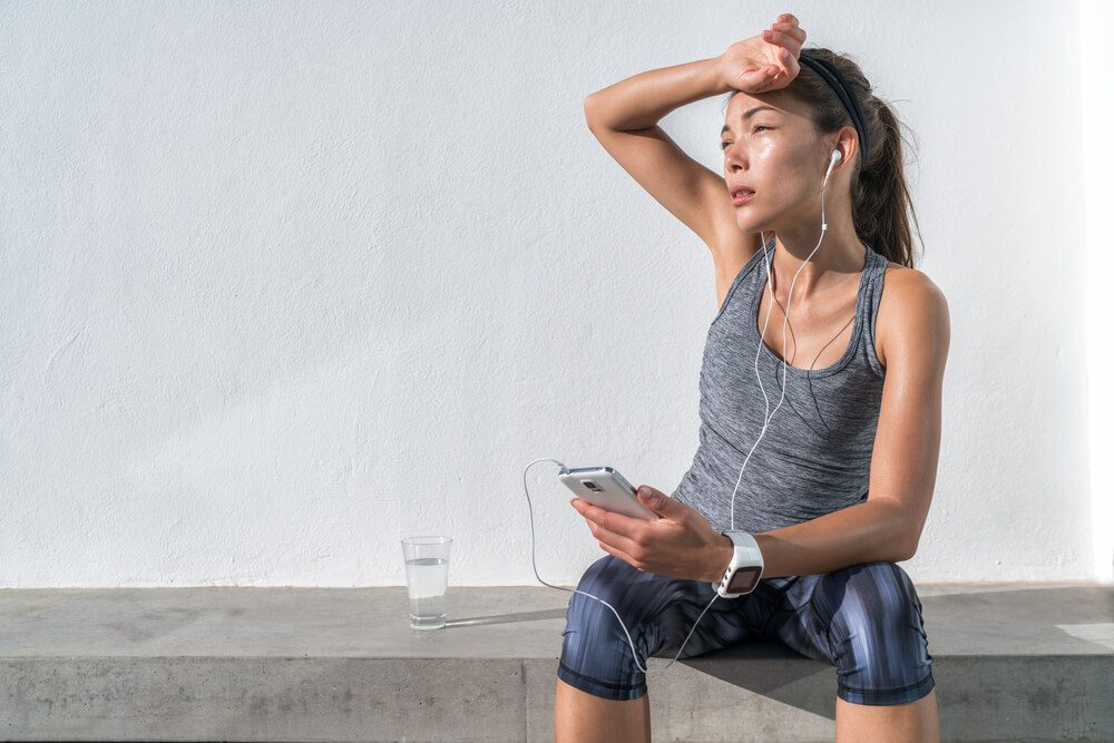 Tired fitness woman sweating taking a break listening to music on phone after difficult training. Exhausted Asian runner dehydrated feeling exhaustion and dehydration from working out at gym.